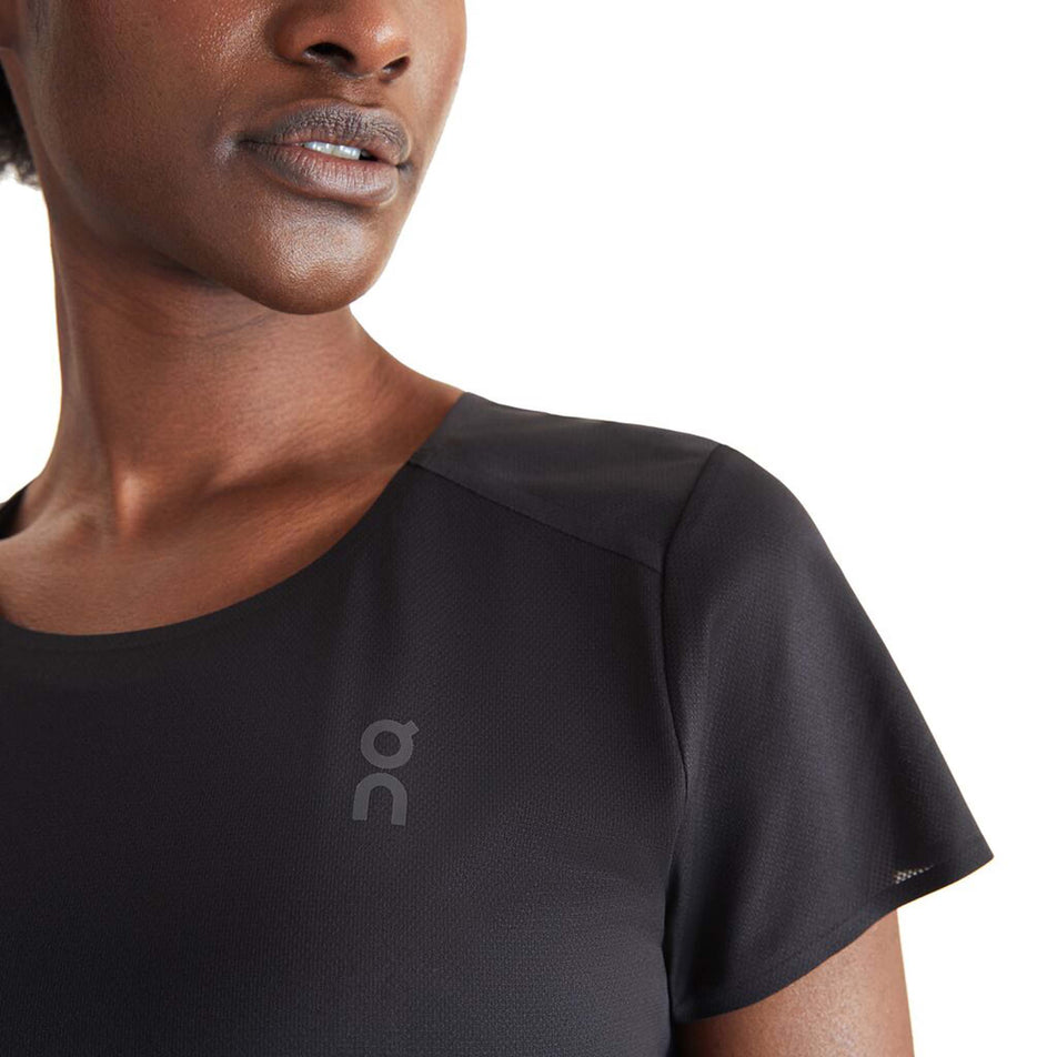 Close-up front view of a model wearing an On Women's Performance T in the Black/Wash colourway. Left side of the upper part of the t-shirt is visible, with the On logo on display. (8002756313250)