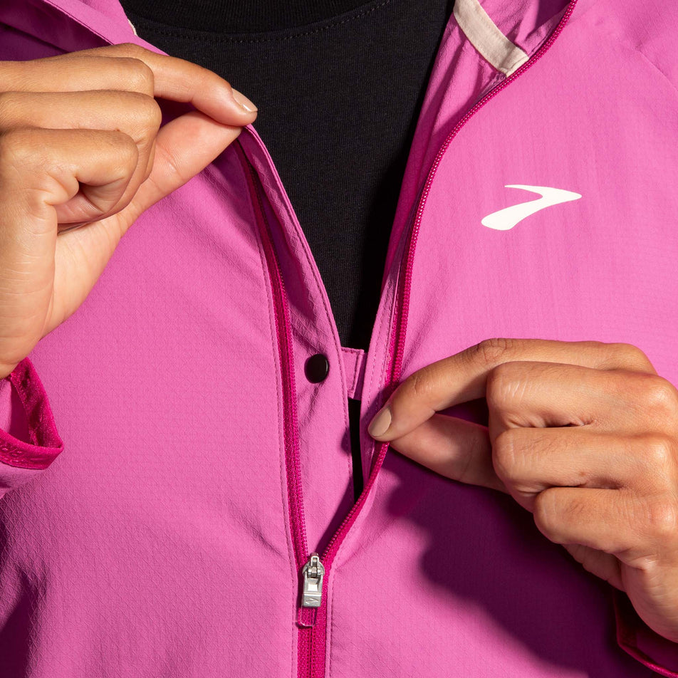 Close-up front view of a model wearing a Brooks Women's Canopy Jacket in the Frosted Mauve/Mauve colourway, demonstrating that the two sides of the jacket can be clipped shut at the chest section when the jacket is unzipped. (8007491748002)