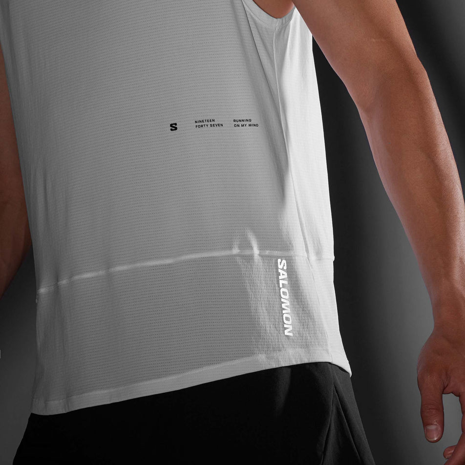 Close-up front view of the logo and brand callout on the front left side of a Salomon Men's Cross Run Tank GFX in the White colourway. Top is being worn by a model in low light conditions. (8246399729826)