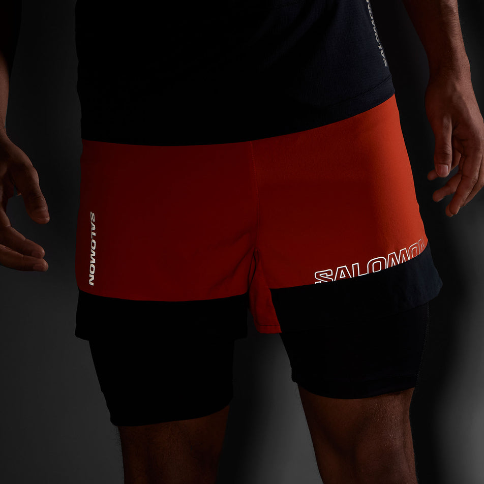 The reflective Salomon logos on the front of a pair of Salomon Men's Cross 2in1 Shorts. Shorts are being worn by a model. (8467295436962)