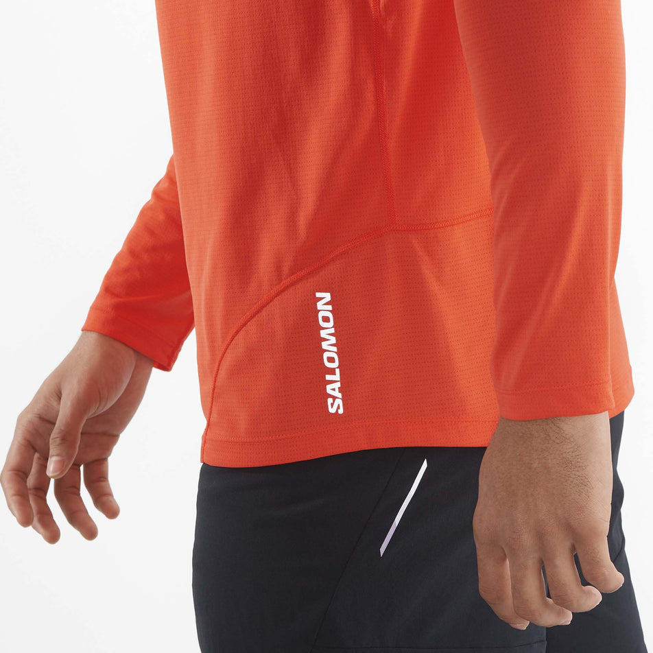 Close-up view of the reflective Salomon logo which is located on the lower front left side of a Salomon Men's Cross Run Long Sleeve T-Shirt. Top is being worn by a model.  (8467287179426)
