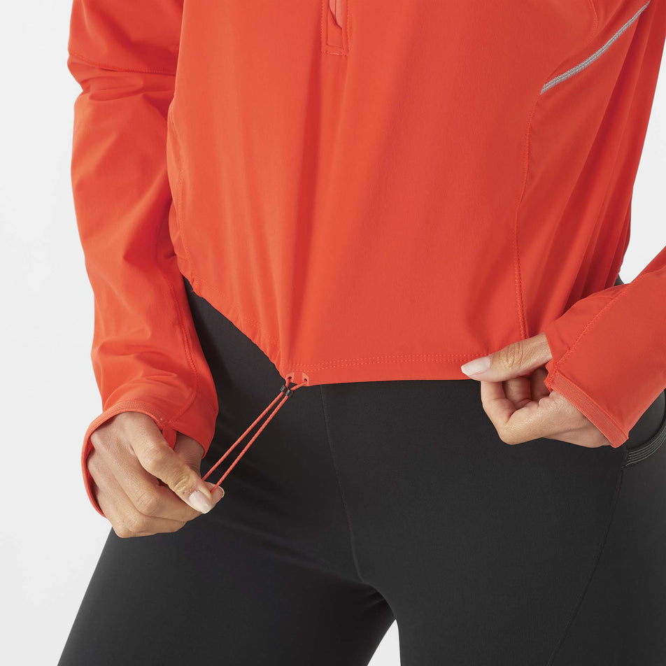 A model highlighting the drawcord on the lower hem of a Salomon Women's Sense Aero Hybrid Cropped Half Zip Midlayer in the Cherry Tomato colourway. Top is being worn by a model who is also wearing leggings.  (8462927102114)