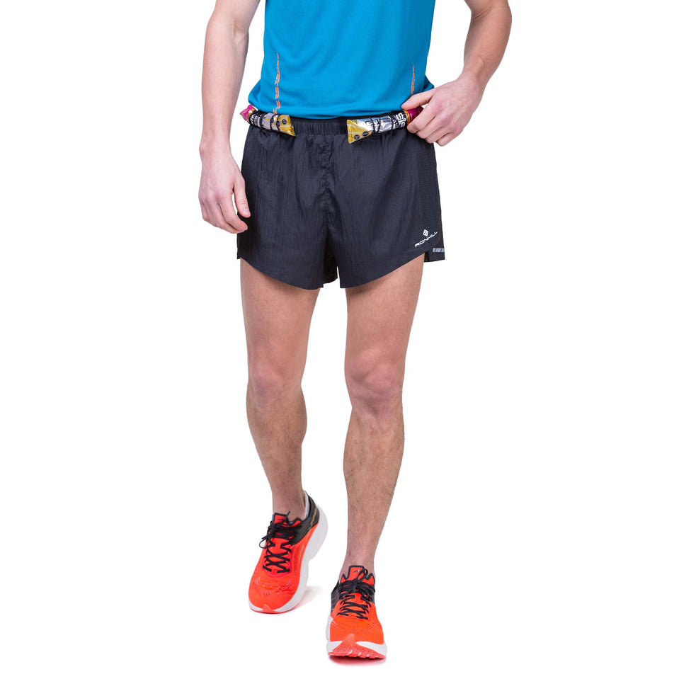 Front view of a model wearing the Ronhill Men's Tech Race Short in All Black colourway, and demonstrating that energy gels can be stored in the elasticated front loops. Model also wearing a Ronhill top and Altra running shoes. (8160873119906)