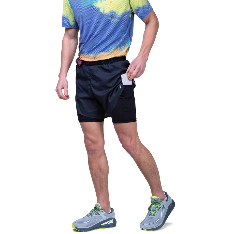Front view of a model wearing the Ronhill Men's Tech Race Twin Short in the All Black colourway. Model demonstrating that a phone can be stored in the stretch pocket on the left side of the inner layer. Model also wearing a Ronhill top and Altra running shoes. (8159263883426)